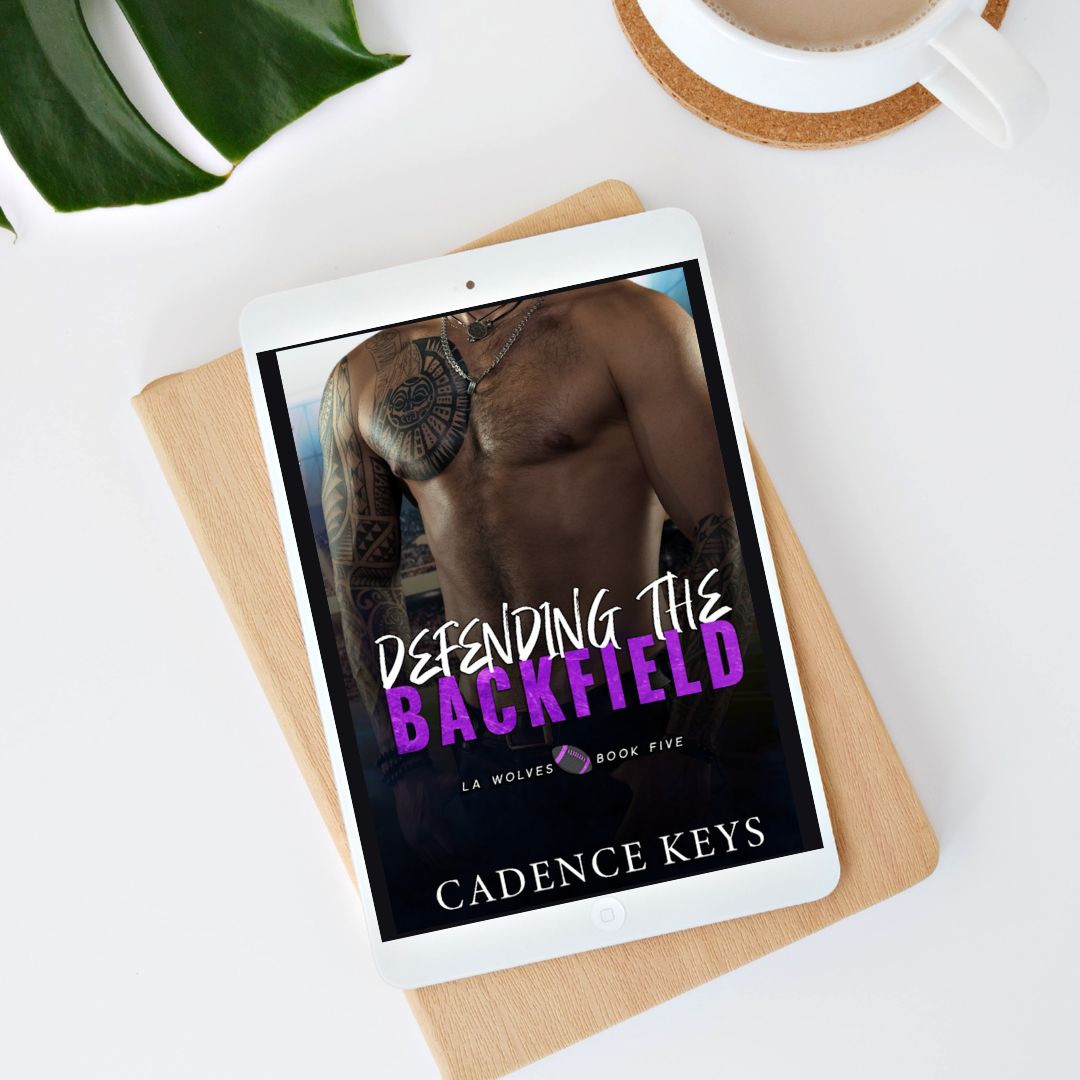 ebook of defending the backfield by cadence keys on top of a white desk with part of a green leaf, a coffee cup, and on top of a book with a wooden cover.