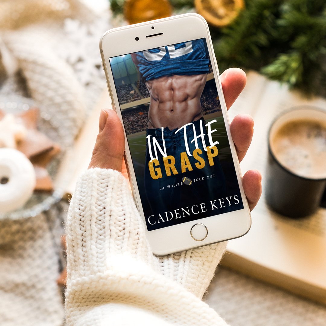 ebook of in the grasp by cadence keys on an iphone held in a woman's hand. Background has blurred visual of a bowl with decorations, a green tree, and a cup of coffee sitting on an open book. 